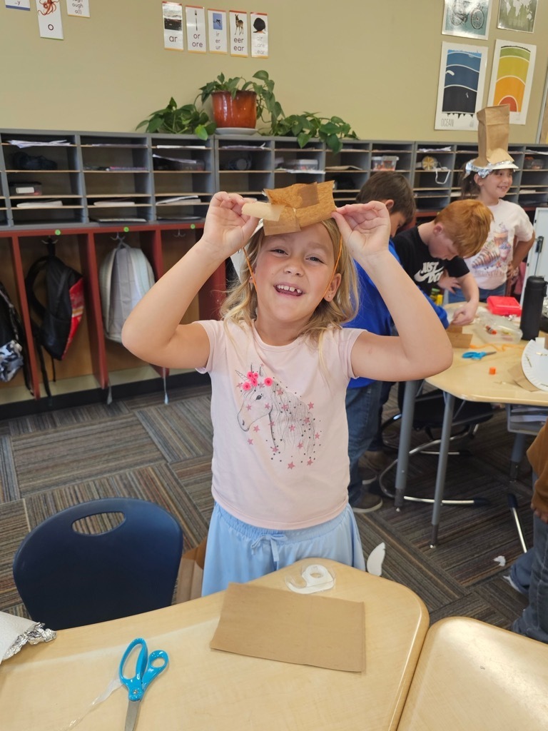 Child holding a cardboard box over head, smiling. Behind, a classroom with tables, chairs, and shelves with plants.