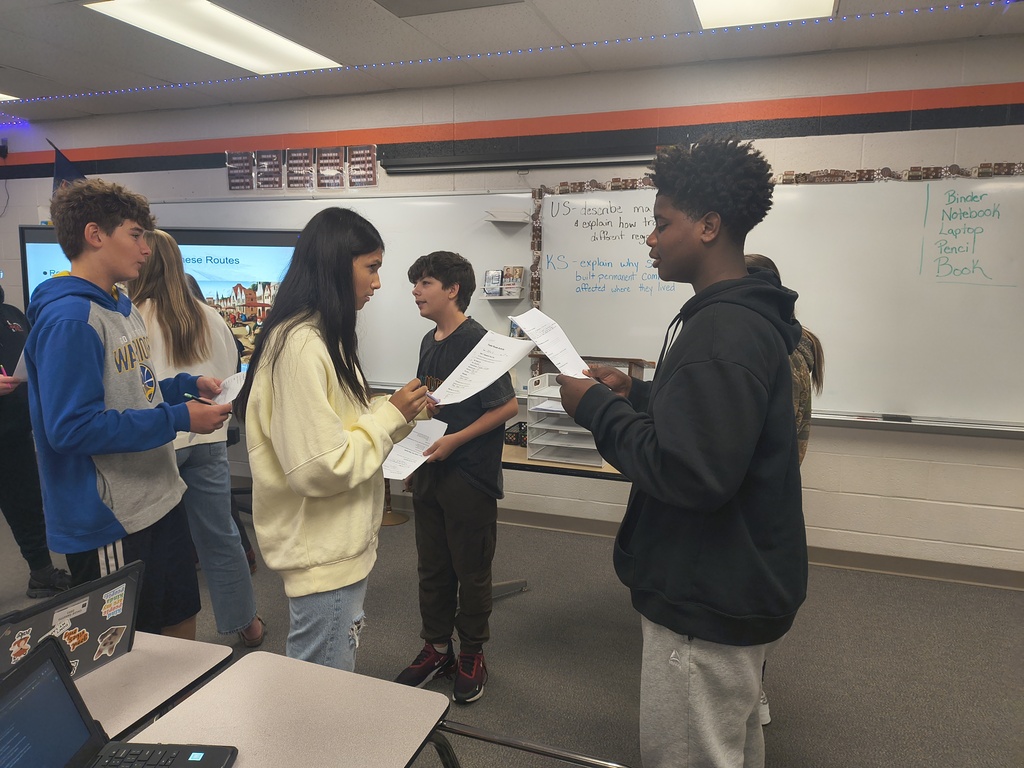 Group of students practicing in a classroom with whiteboards, papers, and laptops on tables.