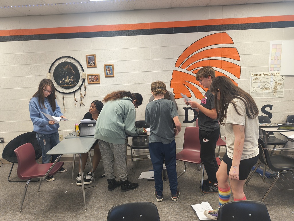 A group of people gather around tables in a classroom. They are looking at papers on the tables.