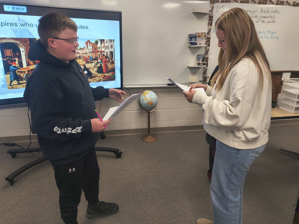 Two students facing each other in a classroom, one holding a paper and the other reading from a paper.