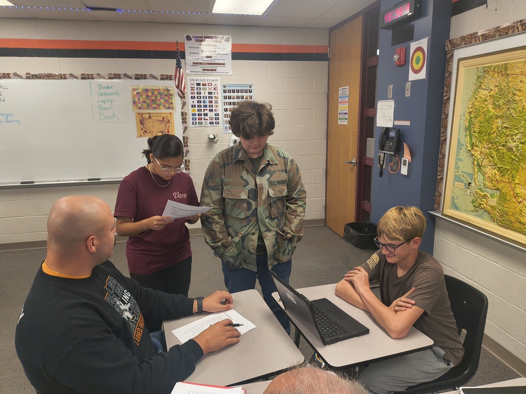 Four individuals are gathered around a table with a laptop, papers, and a map on the wall.