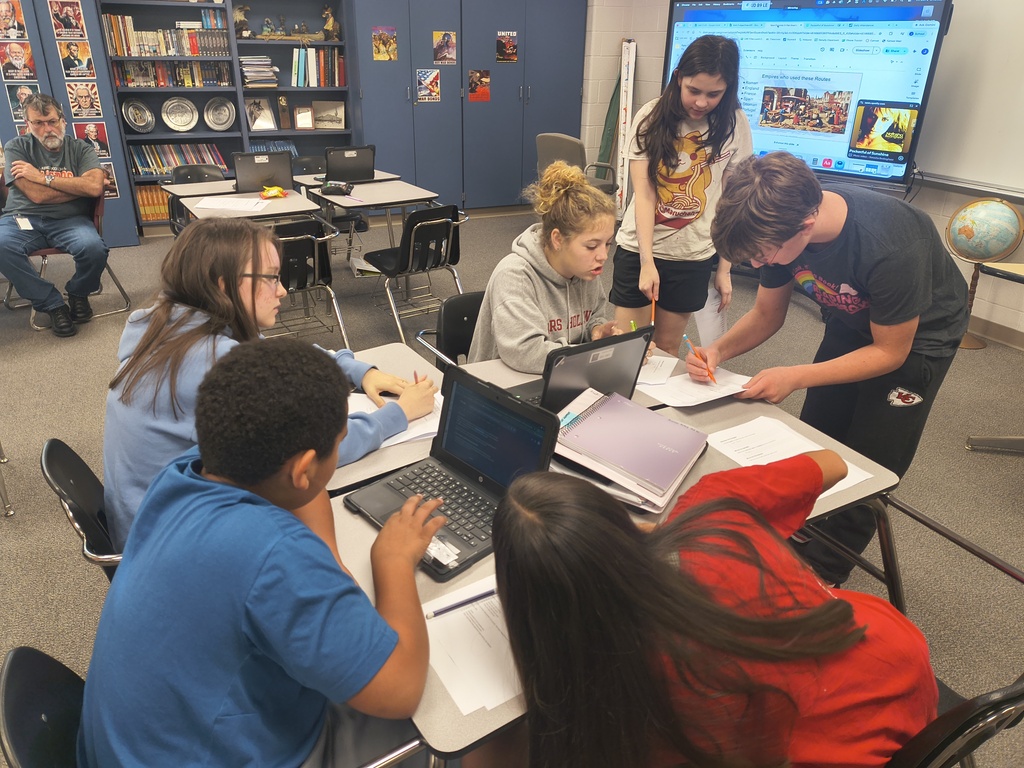 A group of students using laptops and books at tables in a classroom. A teacher stands nearby.