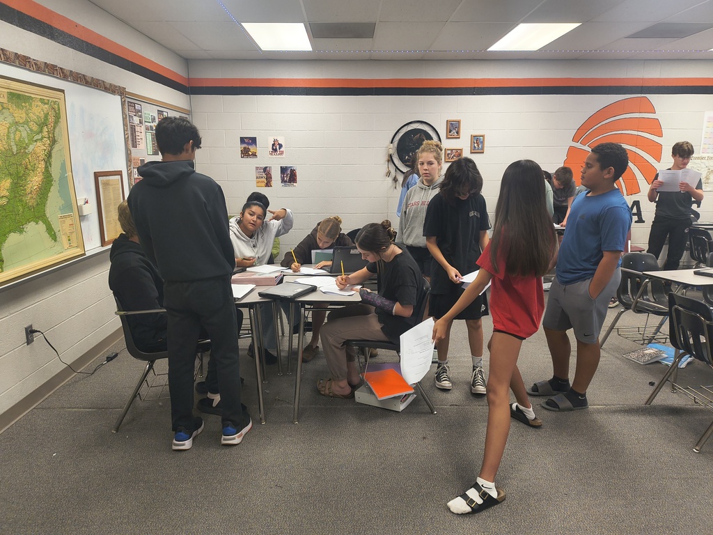 A classroom with a table, chairs, and students working on papers. A large map hangs on the wall.