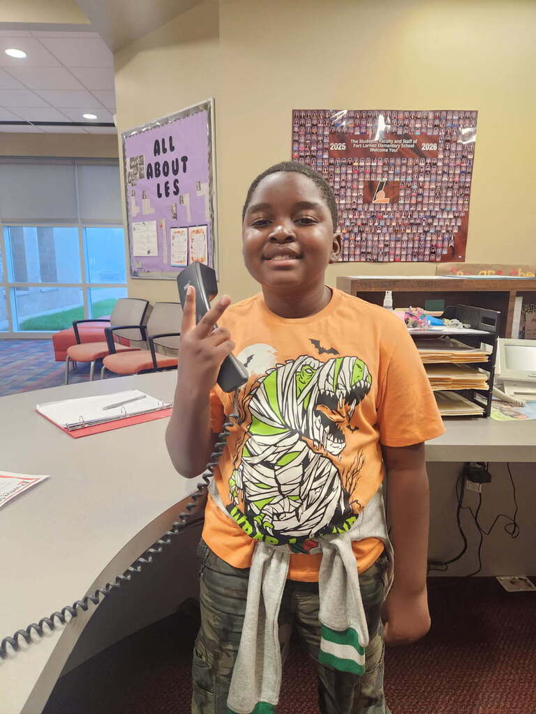 A boy holds a phone in front of his face in an office. Behind him, a desk has a poster.