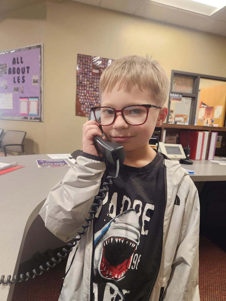 A child holds a phone to their ear. Behind them is a table with a computer, books, and a bulletin board.