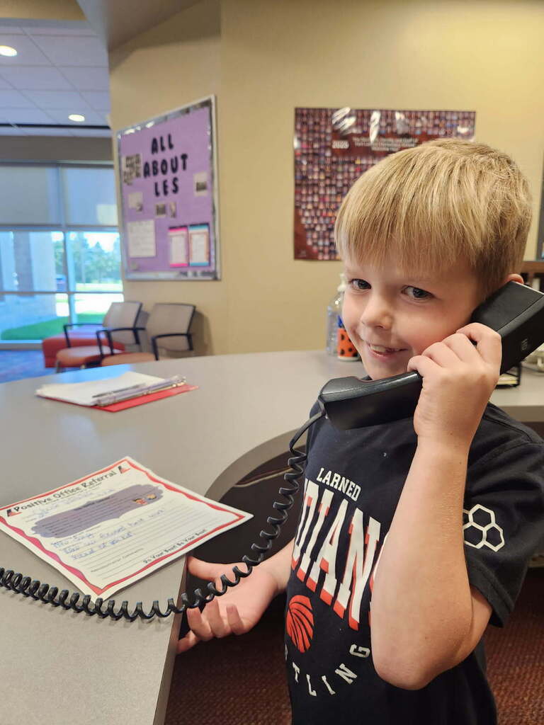 A boy in a black shirt stands at a desk, holding a telephone. Nearby, there is a paper with writing.