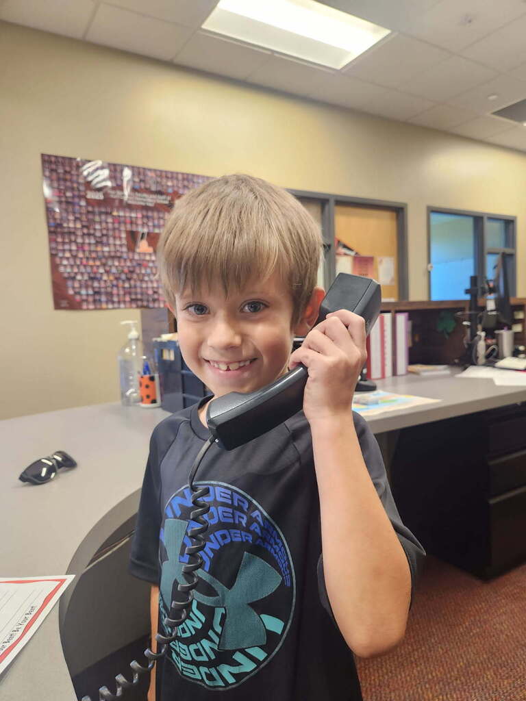 A young boy in a black shirt holds a phone to his ear and smiles in an office.