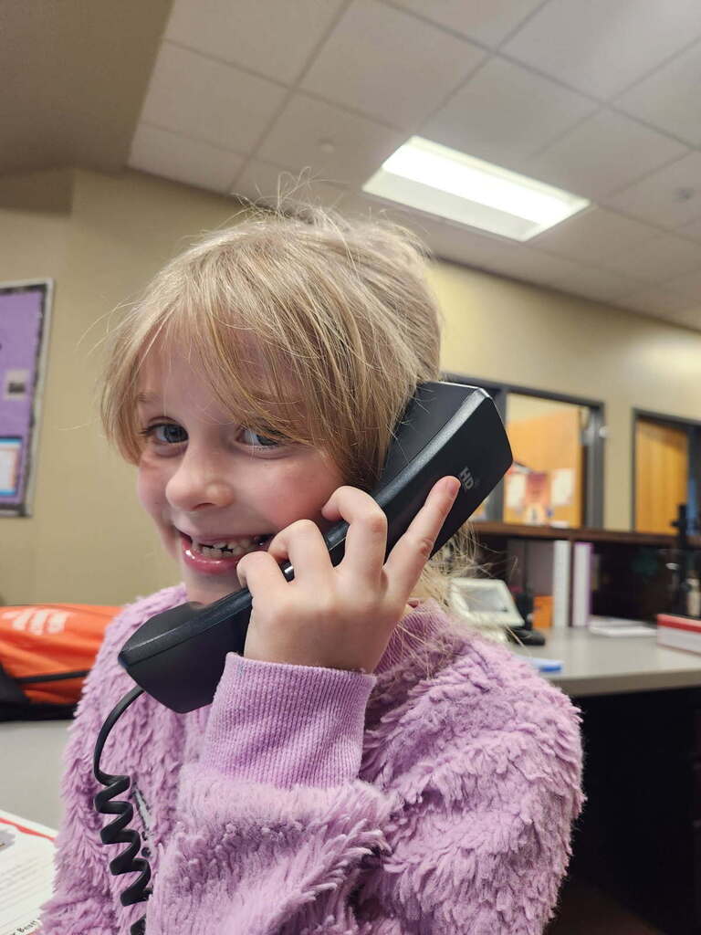 Child wearing pink sweater smiles as they hold a phone to their ear inside a room.