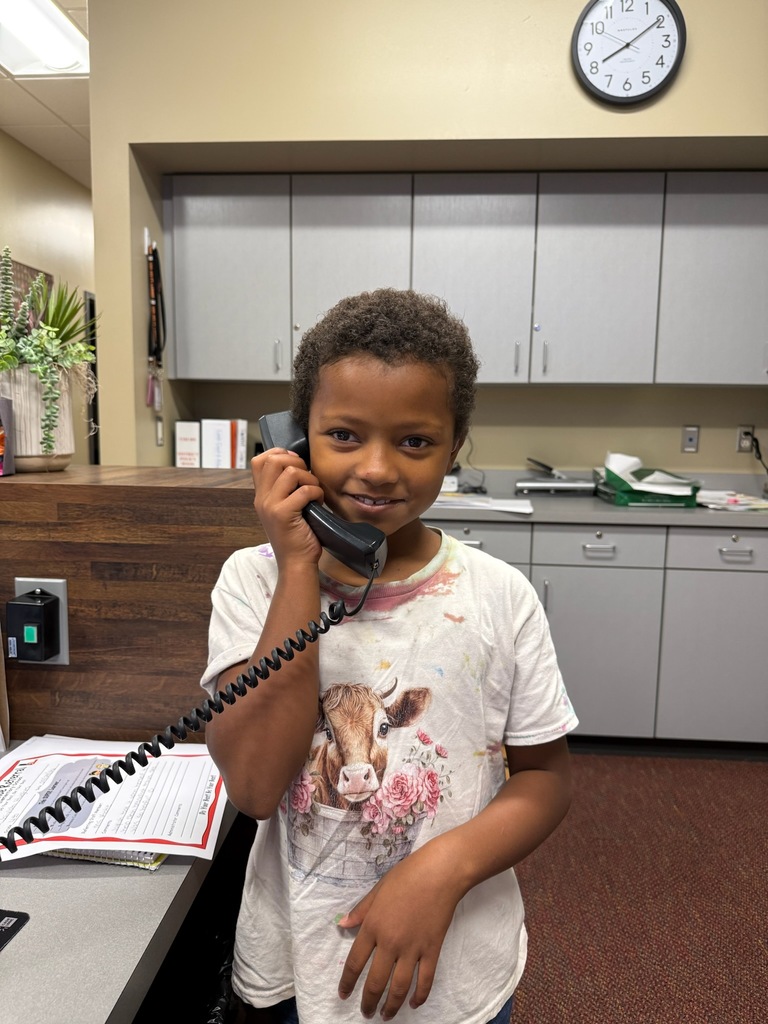 A child with short hair stands and holds a phone, smiling. Behind them, a clock is on a wall.