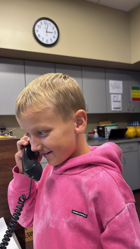 A boy wearing a pink hoodie speaks on a black telephone in a kitchen with cabinets.