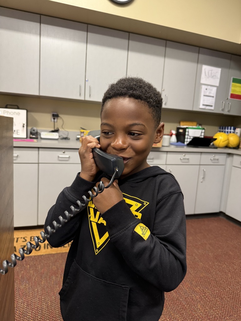 A boy wearing a black hoodie speaks into a phone while standing in a kitchen area.