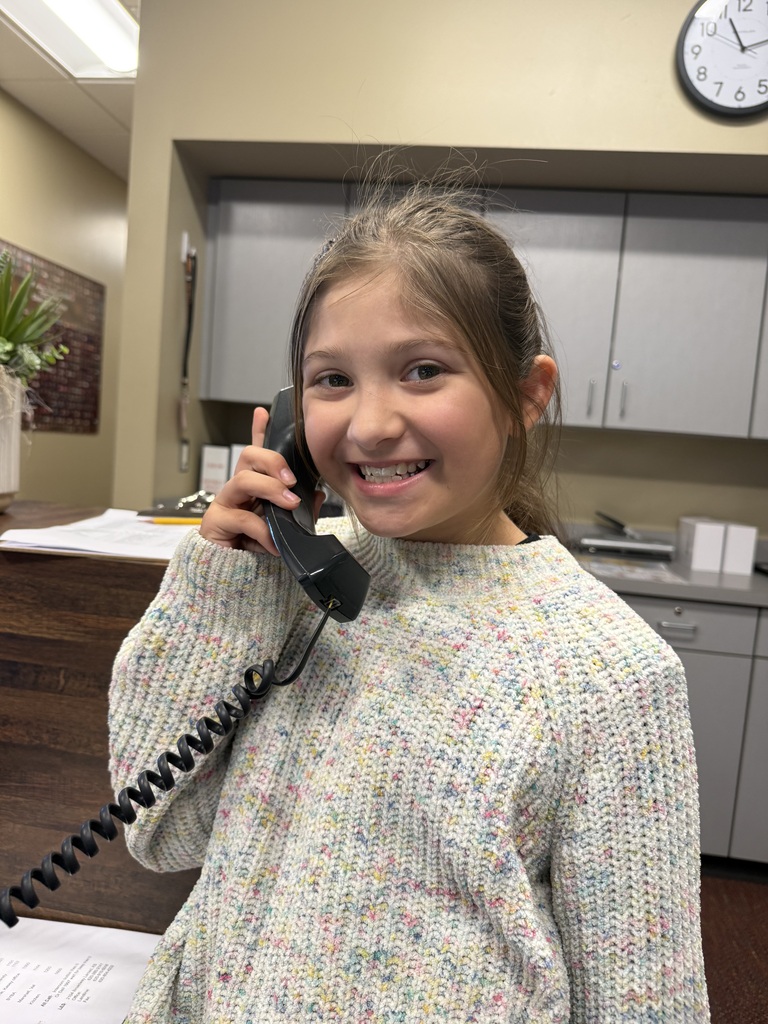 A girl wearing a sweater talks on a telephone inside an office with cabinets and a clock on the wall.