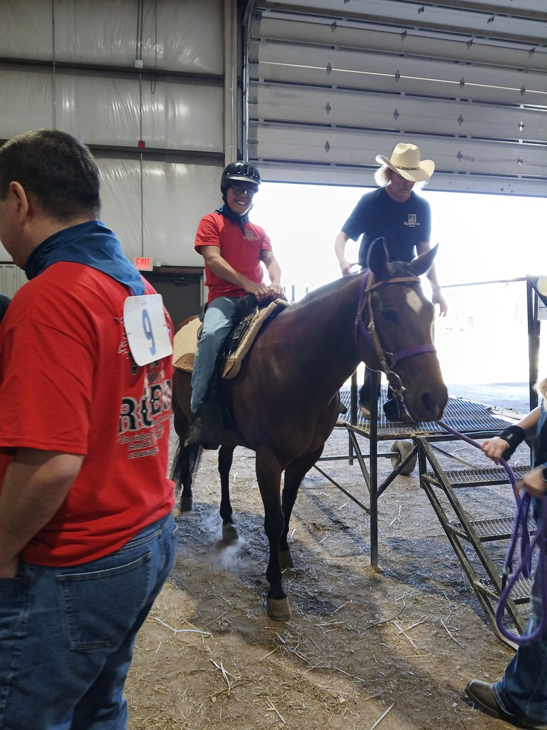 A person in a red shirt rides a brown horse in an indoor space. Two other people are nearby.