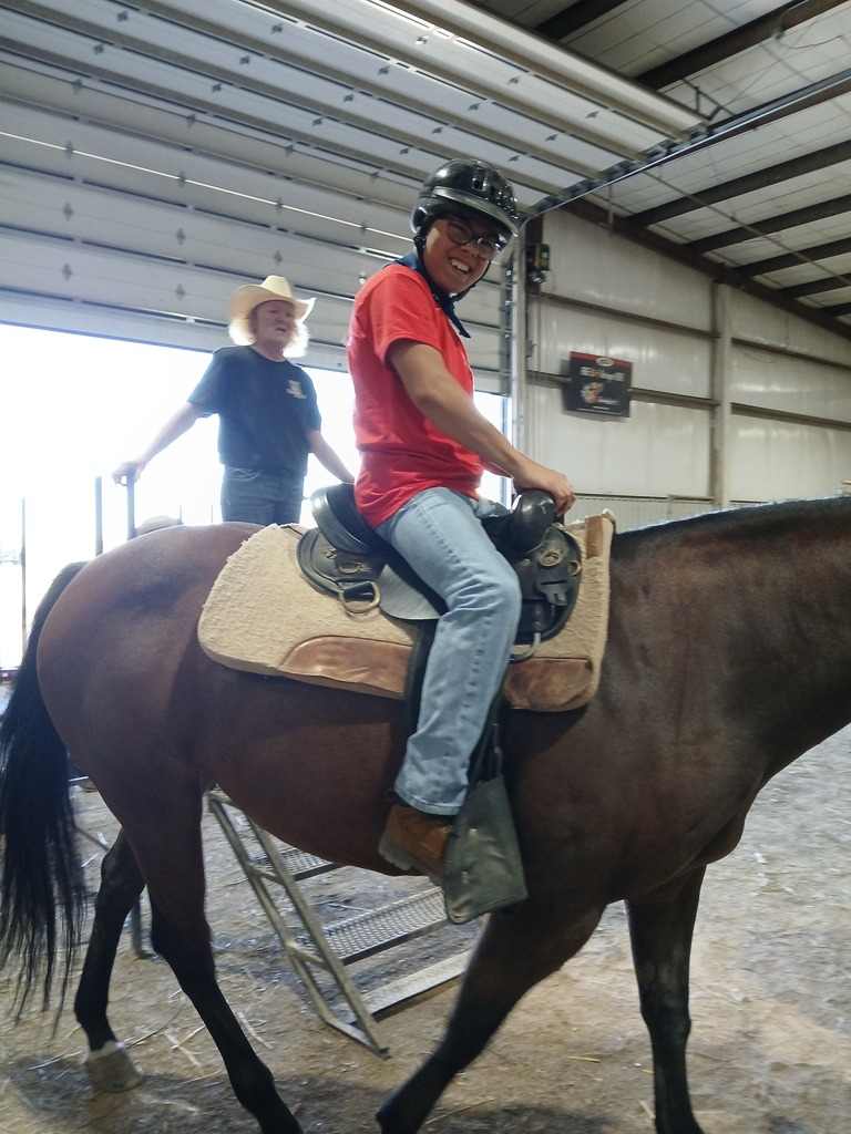 A person in a red shirt and jeans rides a brown horse in a barn. Another person stands behind, wearing a hat and blue shirt.