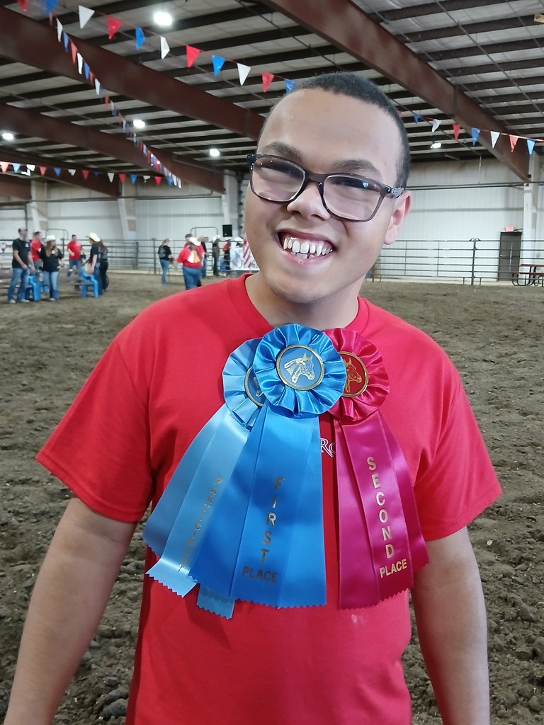 A young person wearing glasses and a red T-shirt stands indoors, smiling. They wear a blue and pink ribbon with text.