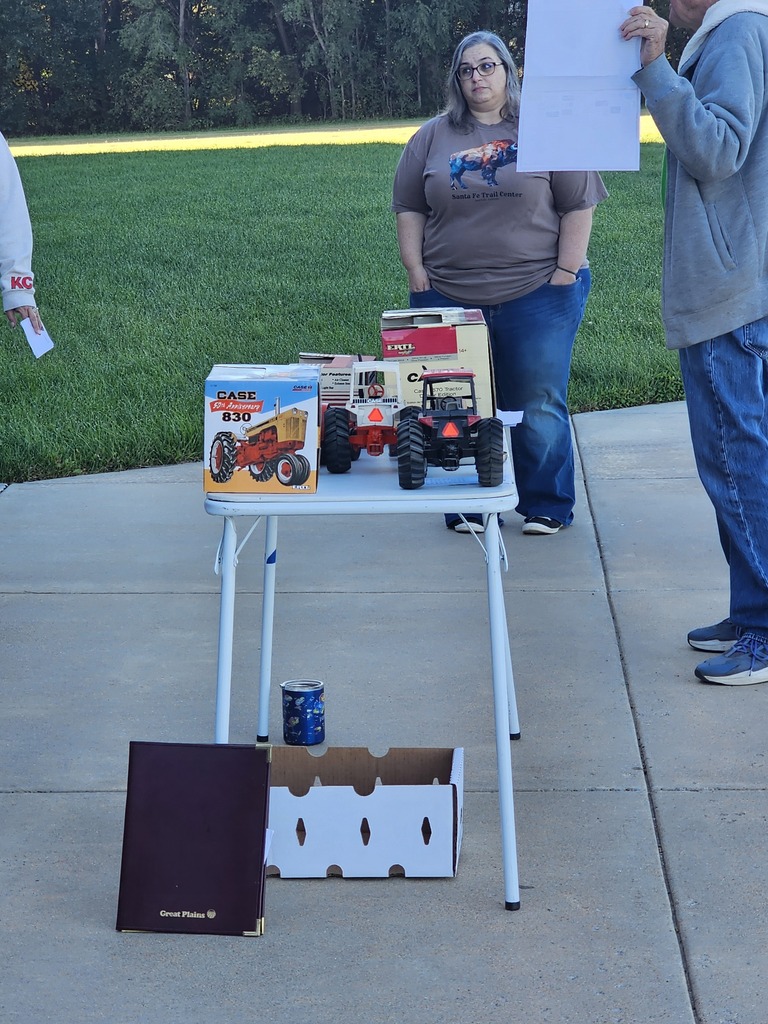 Two people stand by a table with toys and a white paper, set on a driveway with a grassy background.