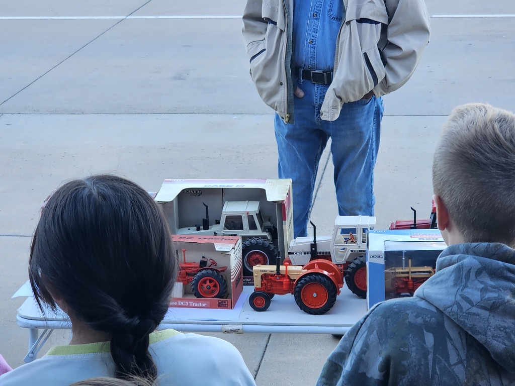 A person stands behind a table with model tractors while two children watch. One child has a ponytail.