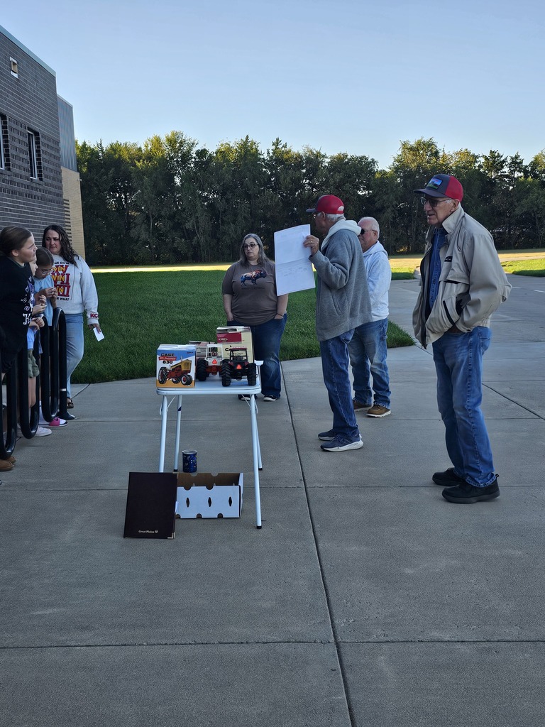 A group of people gather outside near a building. Two men are speaking at a table with items on it.
