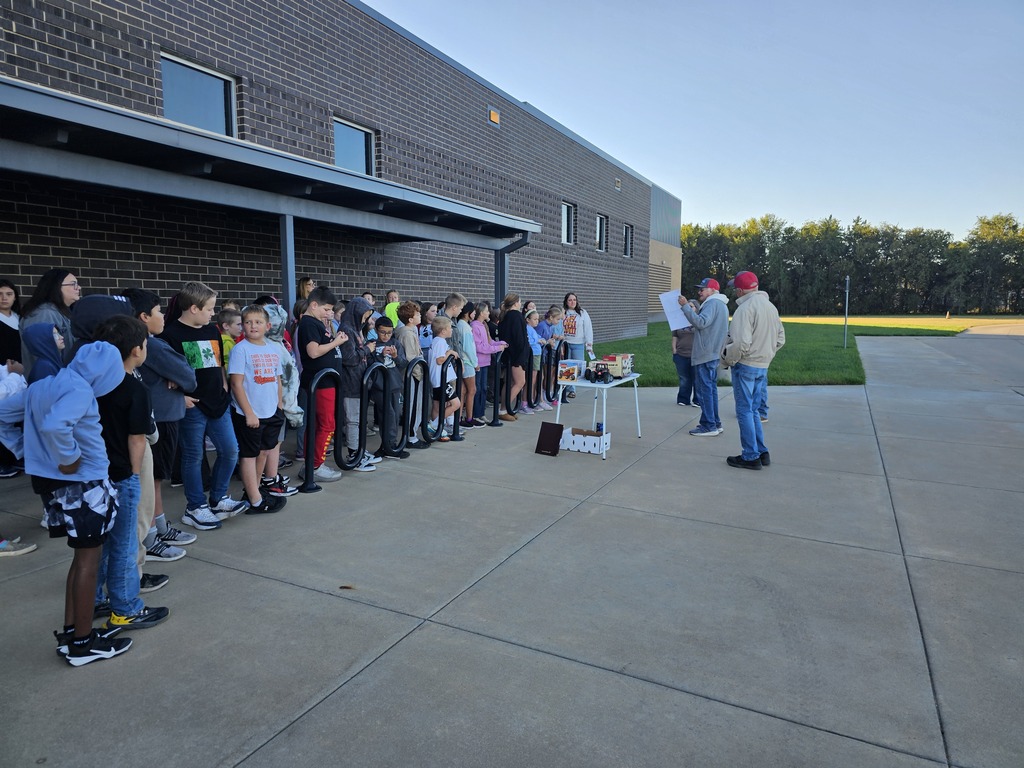 Line of students waiting outside a brick building with a table, two adults, and a grassy area.