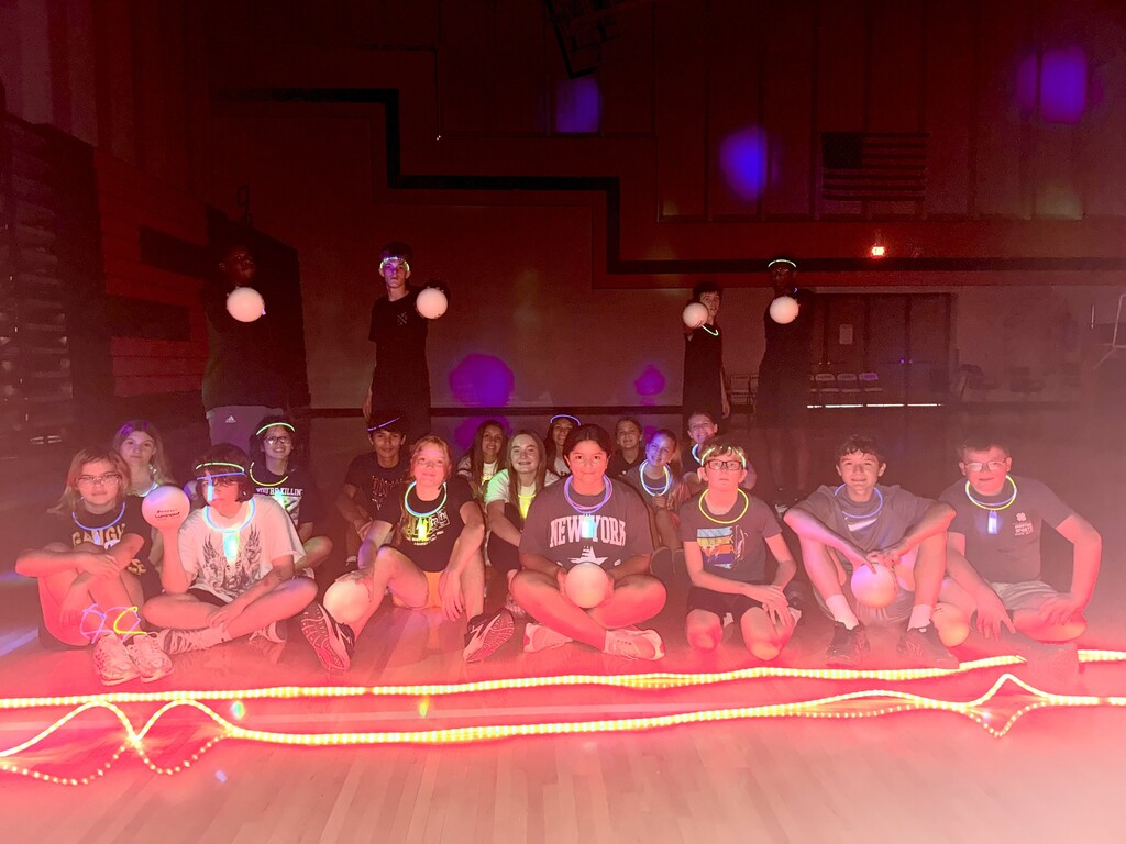 A group of individuals sits on a red platform with colorful lights. They are holding white balls and wearing colorful necklaces.