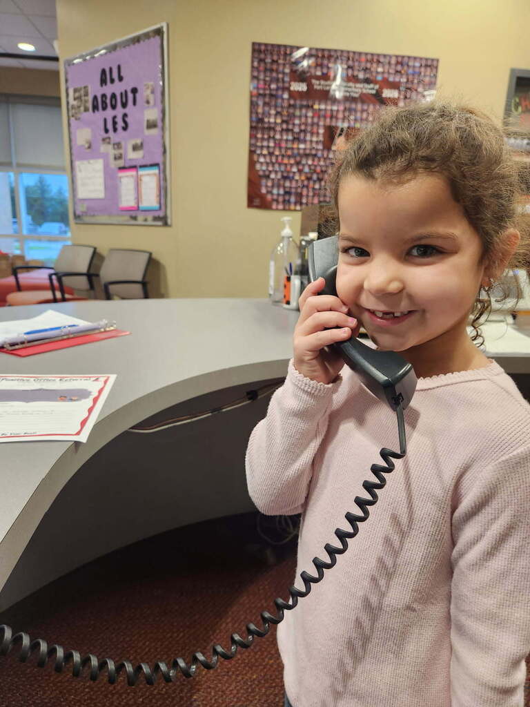 Child wearing pink long-sleeved shirt talking on a telephone with cord, smiling, in an office.