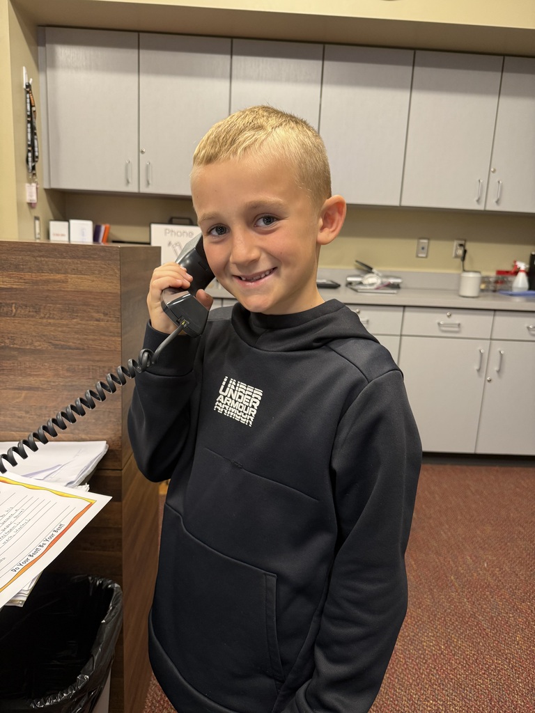Child wearing a dark hoodie, smiling, holds a phone to his ear in a room with cabinets.