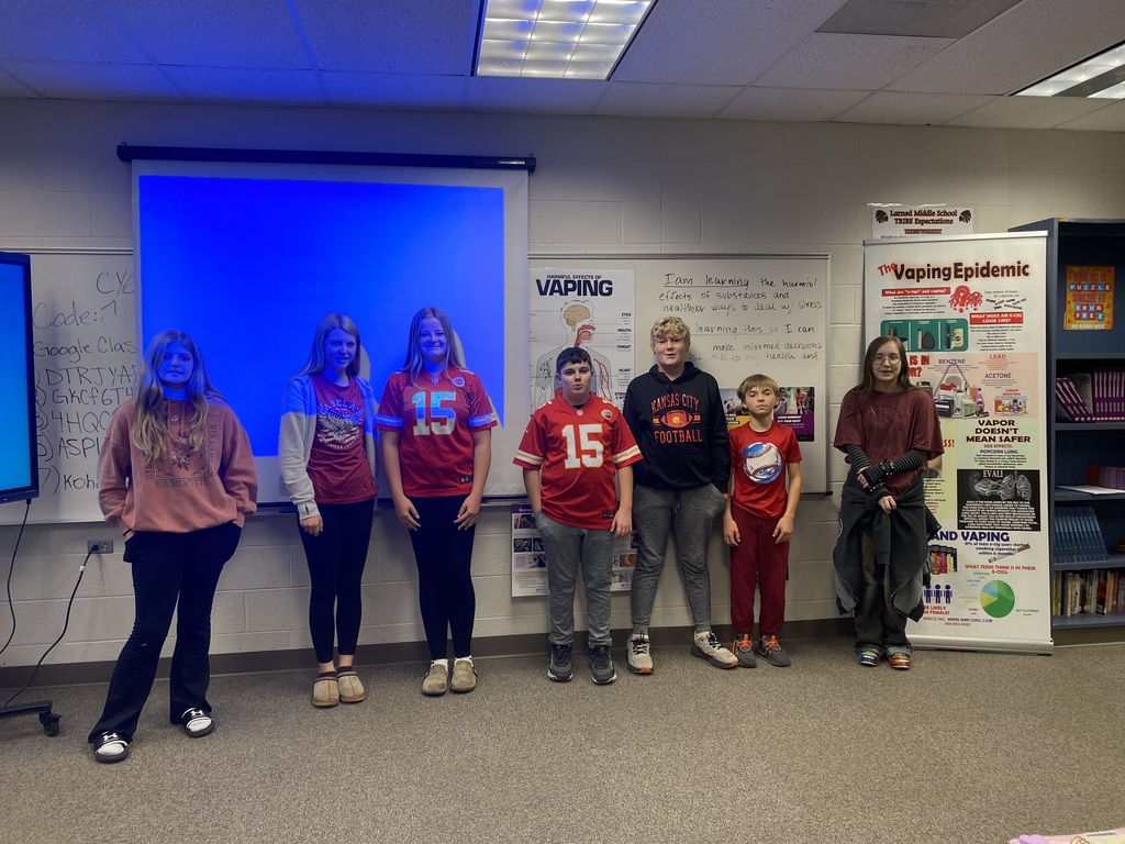 Seven students stand in a classroom, smiling at the camera. A whiteboard and projector screen are behind them.