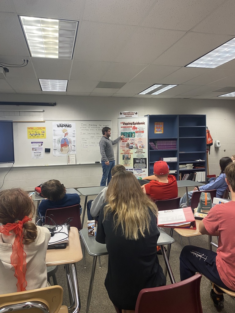 Man stands in front of a whiteboard, gesturing with hand. Several students sit at desks.