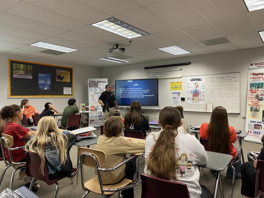 Classroom with students sitting in rows. Teacher standing at front with a screen, pointing at it.