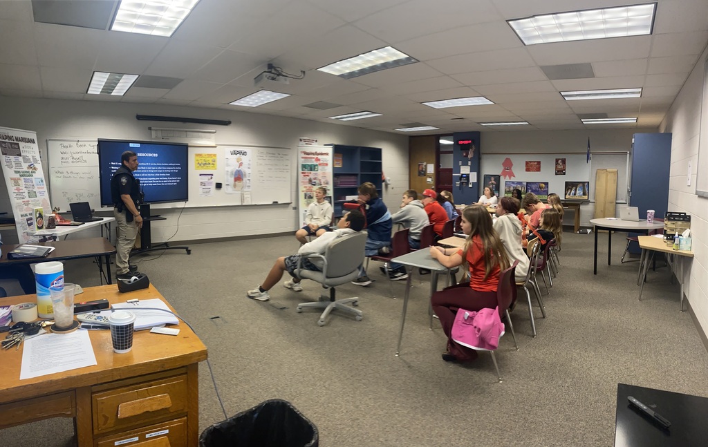 A classroom with a person speaking in front of a screen; students sit in chairs around tables.