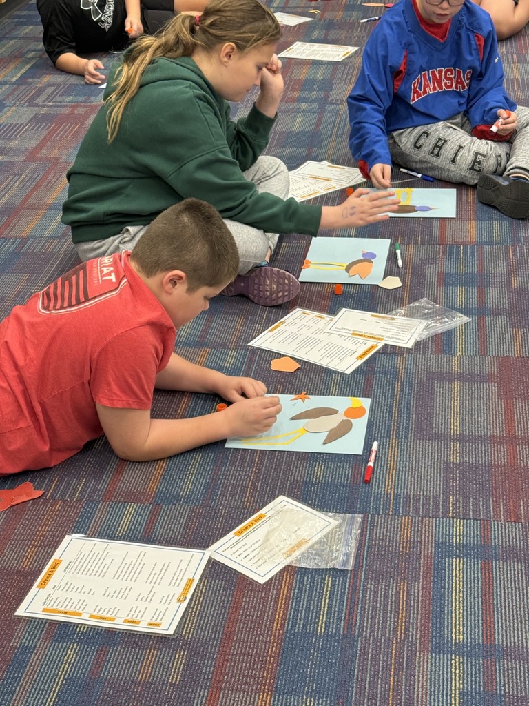 Students sit on the floor, working on papers with crayons and markers. Two boys and one girl are visible.