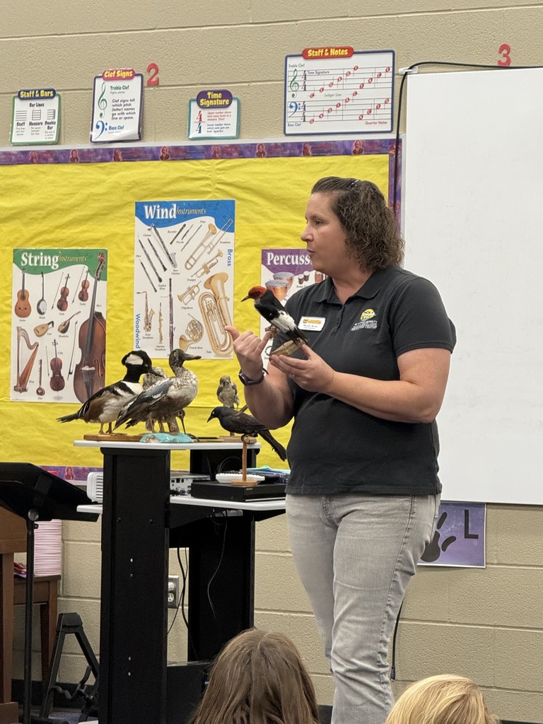 Woman with bird in hand stands at a podium in front of students; several birds sit on podium.