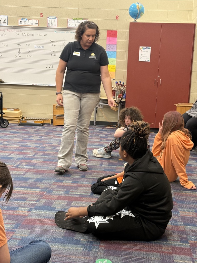 A woman stands before students, some sitting on the floor, in a classroom with a whiteboard and globe.
