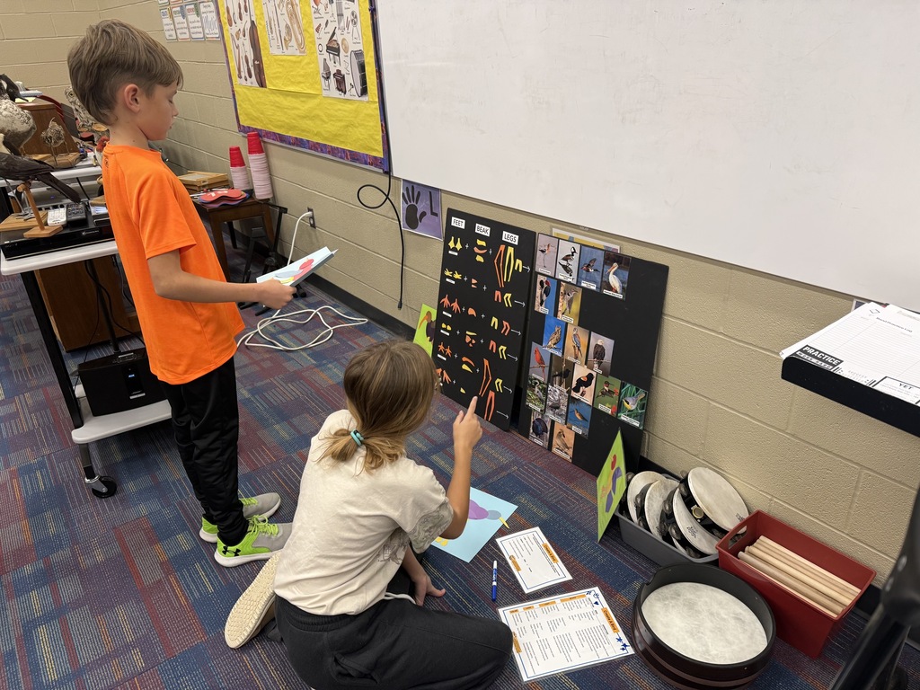 Two children in a classroom, one kneeling on a floor, the other standing, both with musical instruments and papers.