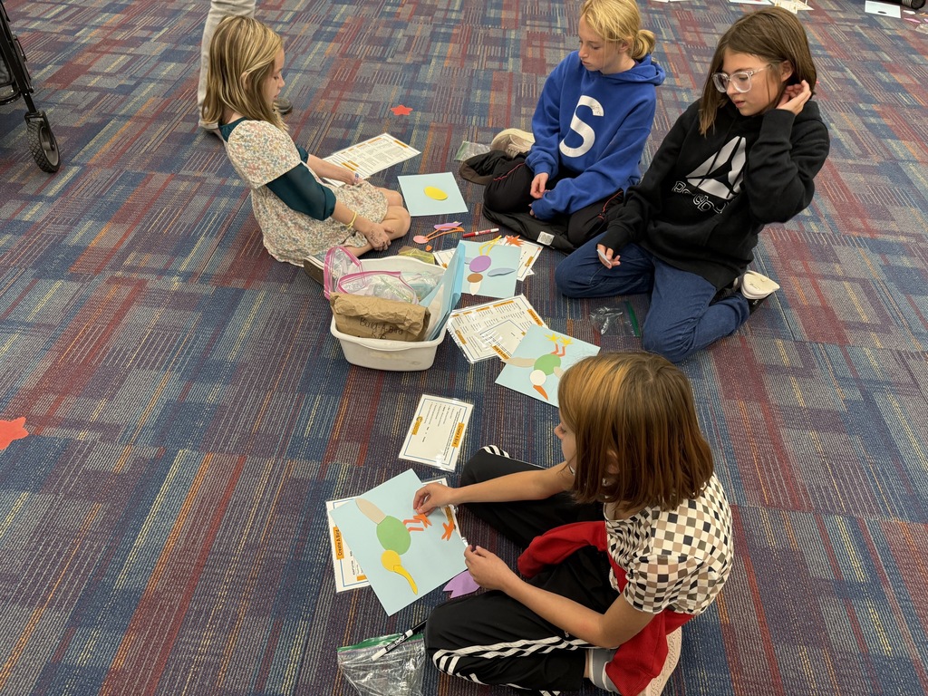 Four children sit on a floor, holding papers, working on crafts. One child holds a small white basket.