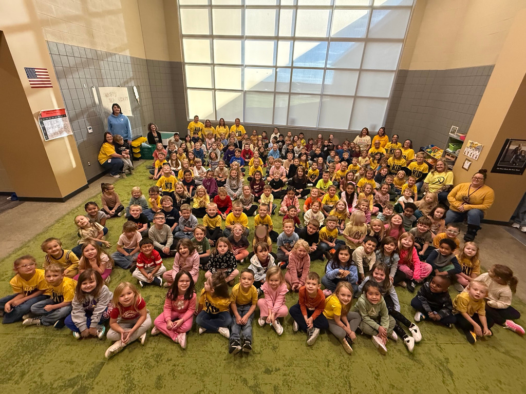 Large group of young students wearing yellow shirts sitting in a circular formation on a green carpet.