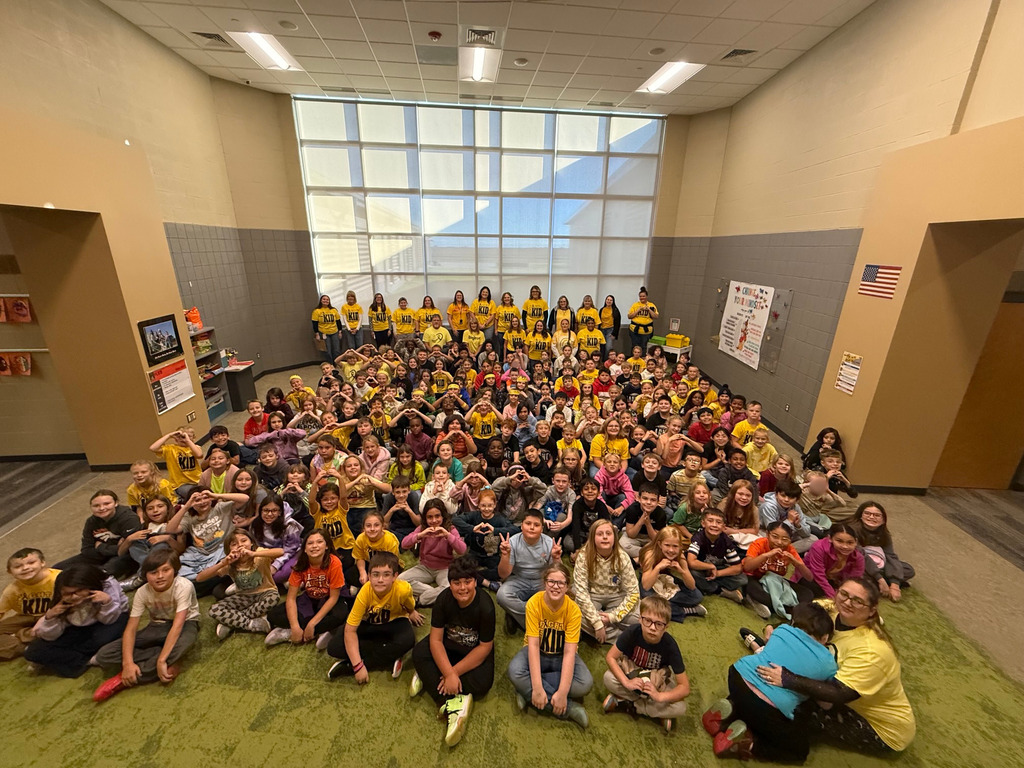 Many students and staff sit on green carpet; some wear yellow shirts; lights on the ceiling.