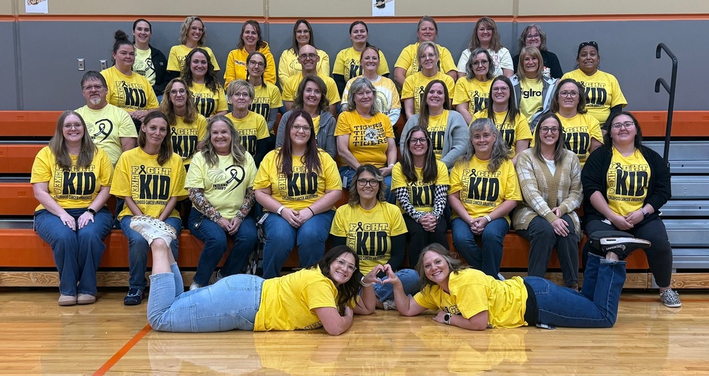 A group of people wearing yellow shirts with KID letters seated on bleachers, with a wooden floor.