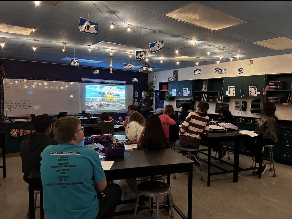 A classroom with students seated around tables, watching a presentation on a screen, with a whiteboard and lights overhead.