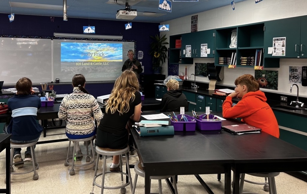 Several students in a classroom with desks and stools, sitting and looking at a screen.