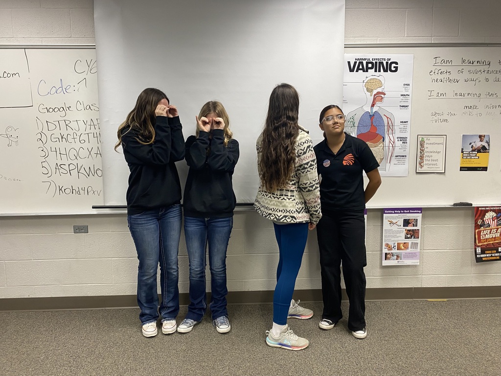 Four girls and a woman stand in a classroom. Three girls cover their eyes with hands. One girl has blue pants.