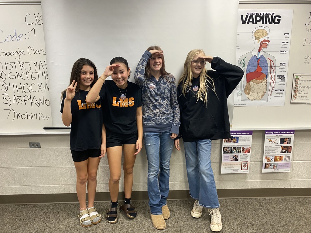 Four young women standing in a classroom, all smiling and posing for a photo. One woman makes a peace sign.