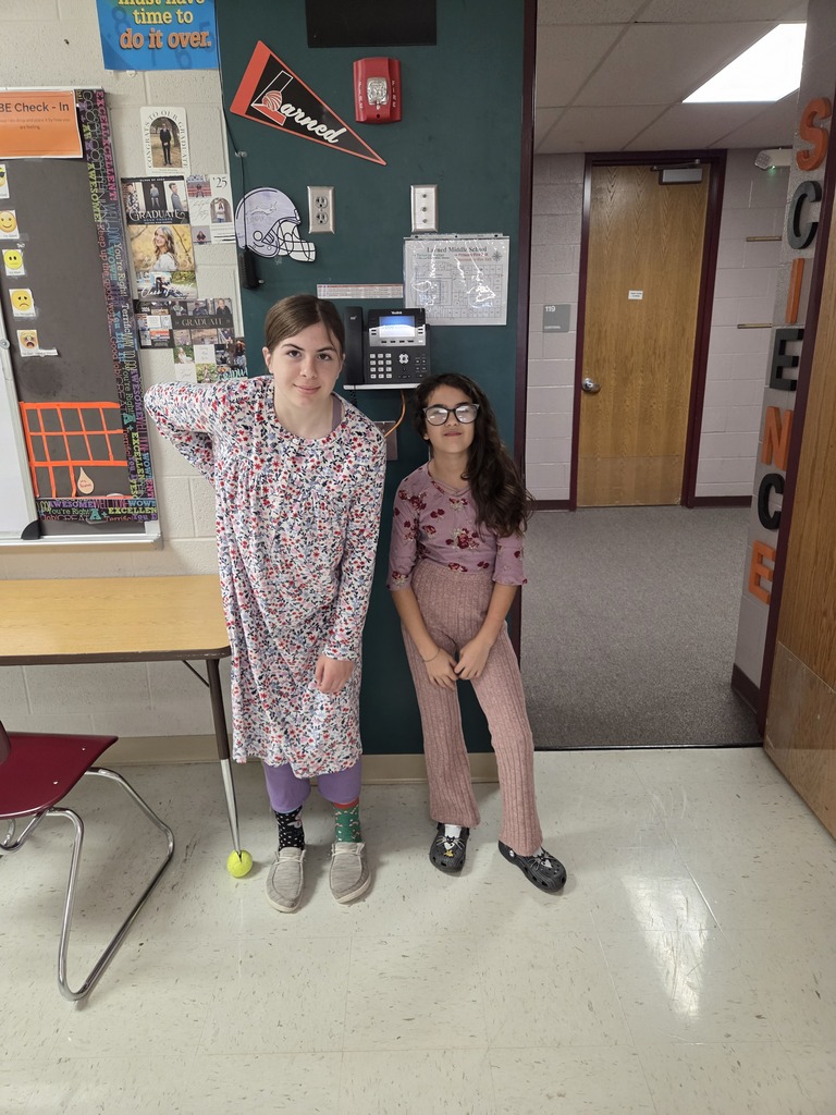 Two students in a hallway, one with long hair and glasses, both smiling at the camera.