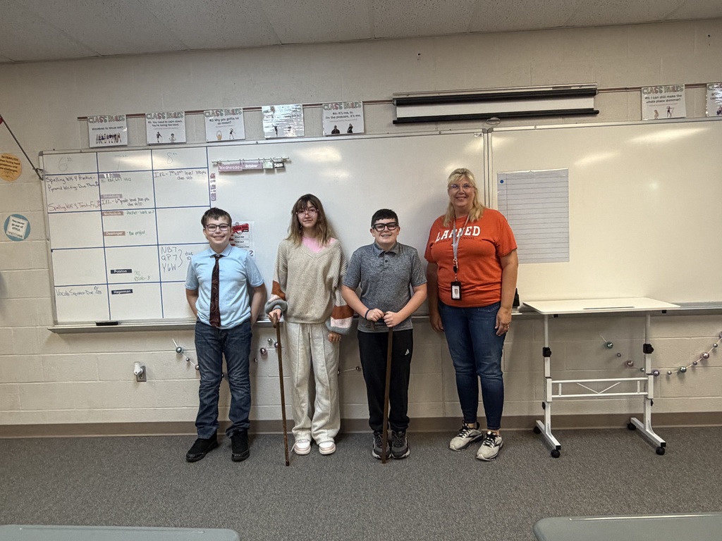Five students stand in front of a white board, one holds a walking stick, another has a tie, and a woman wears a bright orange shirt.