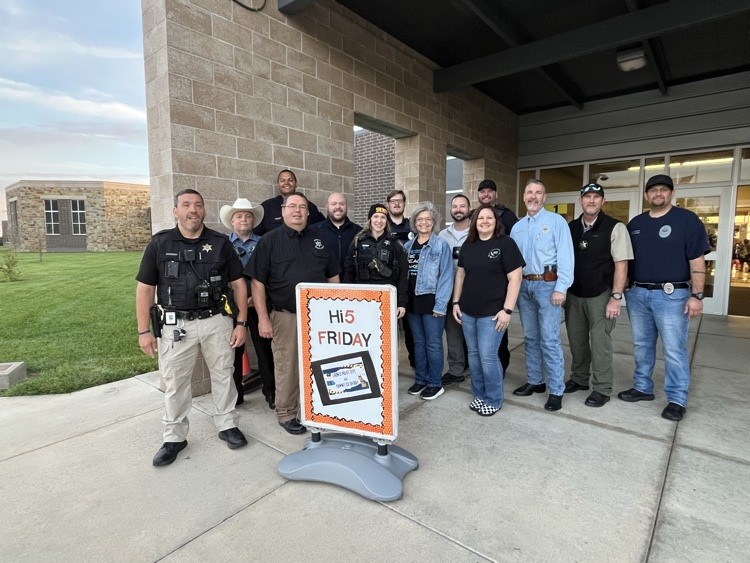 Group of adults in uniform and hats, some with badges, standing by a sign near a building.