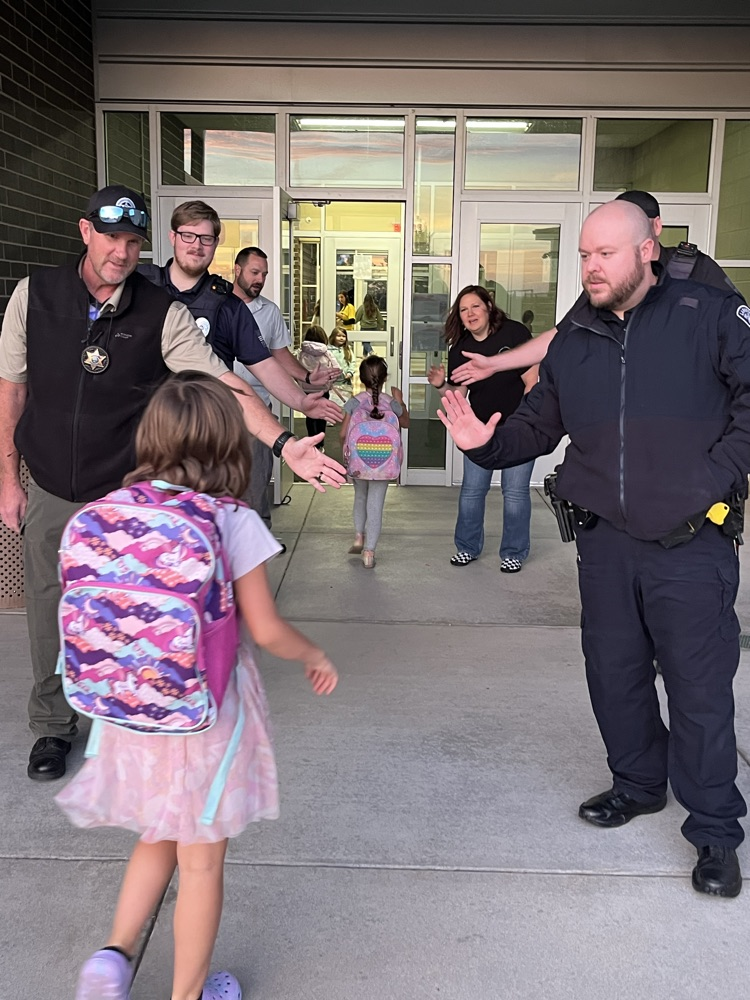 Police officers guide children through the school entrance with hands on their shoulders. One officer holds hands with two girls.