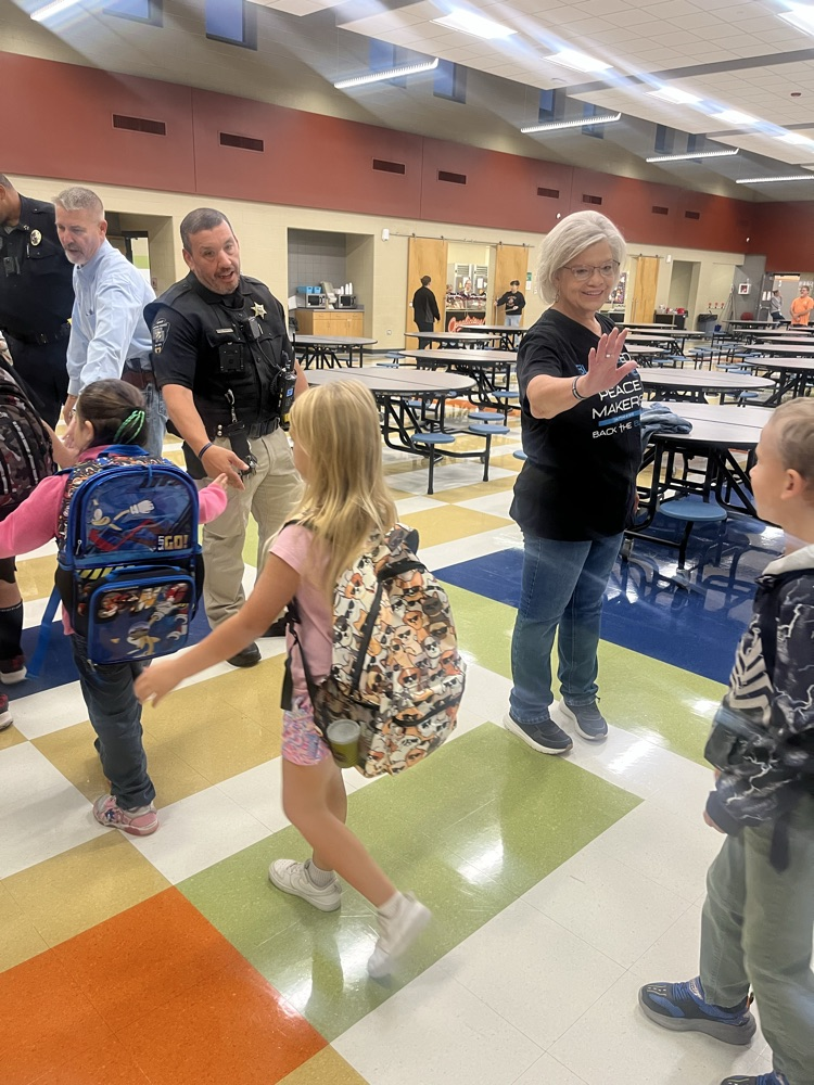 Adults and children with backpacks in a cafeteria with chairs, tables, and a colorful floor.