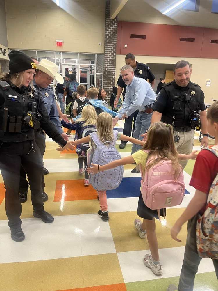Adults, including police officers, greet children in a hallway. Children carry backpacks. Colorful floor squares.
