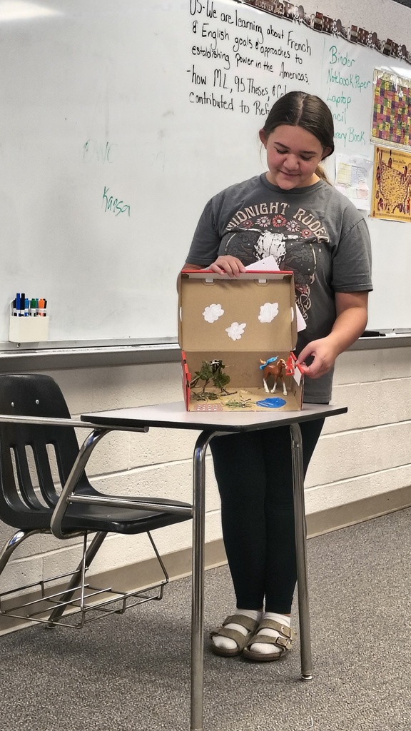Person holding a box with a picture of a farm scene, standing by a table in a classroom.