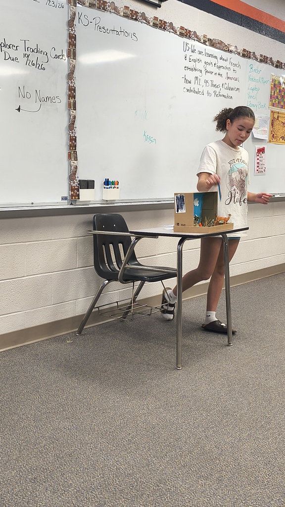 A student stands by a desk holding a small box, near a whiteboard with writing on it.